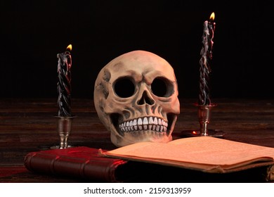 Satanic Composition Of Humans Skull With Satanic Book And Burning Candles. Rithual With Opened Satanic Bible.