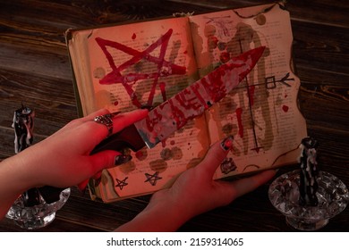Satanic Wicca Ritual Concept. Female Hand Holds Bloody Knife Over The Bible With Pentagram.