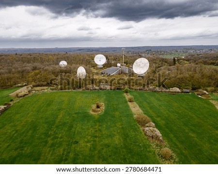 Satellite dish array with control building and antenna tower seen from drone under moody overcast sky, telecommunications ground station infrastructure amid green farmland