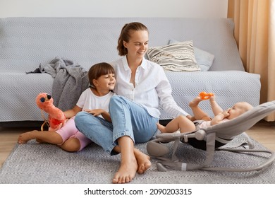 Satisfied Adorable Woman Wearing White Shirt And Jeans Sitting On Floor Near Cough, Looking At Baby In Bouncer With Smile And Great Love, Spending Time With Her Children.