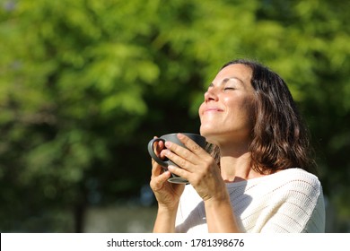 Satisfied adult woman smelling coffee cup standing in a green park at summer