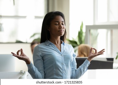 Satisfied African American Businesswoman Relaxing, Meditating In Yoga Pose Breathing Exercises At Workplace. Mindful Diverse Employee Standing With Eyes Closed At Office, Stress Relief Concept.