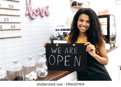 Satisfied African American female barista or waitress, owner of a coffee shop or restaurant, stands holding a sign we are open near the bar counter and friendly smiling - Powered by Shutterstock