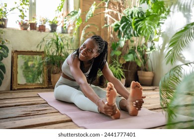 Satisfied african american woman enjoy fitness time sit on yoga mat stretching with smile surrounded by tropical indoor plants. Happy sported black female do exercises for flexibility and healthy body - Powered by Shutterstock