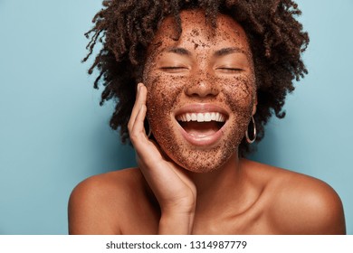 Satisfied African American woman has pore cleaning with scrub mask, laughs happily and shows white teeth, has curly hair, healthy skin, isolated over blue studio wall, uses peelig cosmetic product