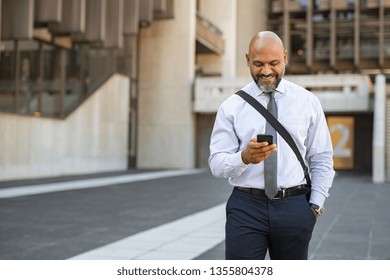 Satisfied African Businessman Using Smartphone While Walking In The Street. Mature Smiling Business Man Walking On The Road And Messaging With Phone. Happy Successful Entrepreneur Going To Work.