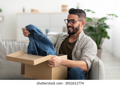 Satisfied Arab male client taking out new clothes from cardboard box, happy with delivery service at home. Millennial Eastern guy unpacking cardboard parcel, receiving online store order, indoors - Powered by Shutterstock