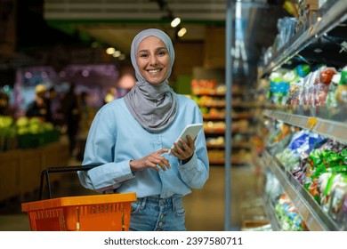 Satisfied Arab woman in hijab is shopping in the store, holding a phone and a shopping basket, smiling and looking at the camera. - Powered by Shutterstock