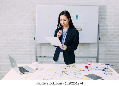 Satisfied Asian Woman In Formal Outfit With Badge Smiling And Reading Paper While Standing By Table With Marketing Plan And Laptop In Office