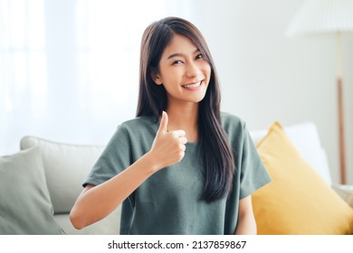 Satisfied asian woman looking at camera with thumbs up sitting on a couch in the living room at home. - Shutterstock ID 2137859867