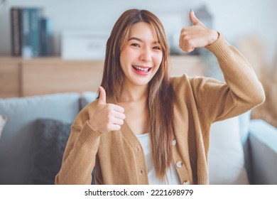 Satisfied asian woman smiling while looking at camera with thumbs up sign sitting on a couch in the living room at home. Body language. I like that
