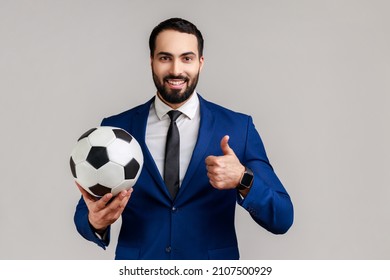 Satisfied bearded businessman holding soccer ball on his hand with smiling positive expression, showing thumb up, wearing official style suit. Indoor studio shot isolated on gray background.
