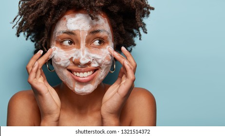 Satisfied beautiful black woman makes massage of face, has white foam, problem of wide pores, cares of skin, keeps gaze aside, smiles positively, models over blue studio wall with free space