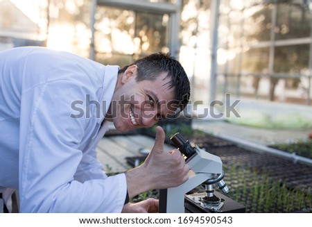 Satisfied biologist looking through microscope and showing thumb up in greenhouse with sprouts