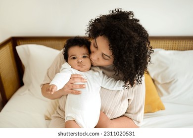 Satisfied black millennial mom kiss little baby, sit on white bed, enjoy love, comfort and rest in bedroom interior. Kid care vacation, parenthood, health care and family at home, ad and offer - Shutterstock ID 2311020531