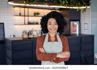 Satisfied black woman in apron standing with crossed arms on chest on background of bar counter. Confident small business owner in service sector ready for accepting clients in beginning of day. - Powered by Shutterstock