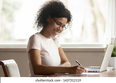 Satisfied Black Woman Housewife In Glasses Sitting At Table Near Laptop Holding Pen Writing Planning Family Budget Making Notes, Businesswoman Working At Home, Girl Studying Online Noting Information