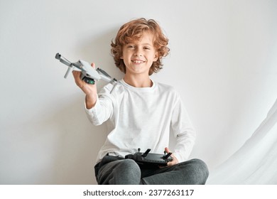 Satisfied blond child with toy drone in raised hand looking at camera against white wall - Powered by Shutterstock