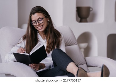 Satisfied Brunette Businesswoman In Glasses, Shirt Black Pants Sitting On Cozy Chair With Opened Planner Smiling Broadly, Checking To Do List. Pretty Hispanic Young Woman At Home Preparing For A Day.