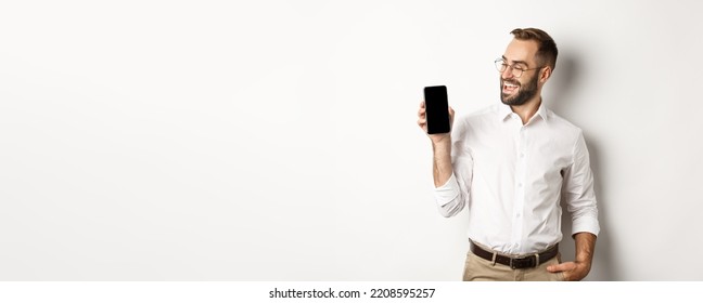 Satisfied Business Man Showing And Looking At Mobile Screen, Introduce Application Or Website Promo, Standing Over White Background