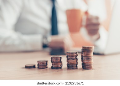 Satisfied businessman drinking coffee with coin stack in front, close up with selective focus - Shutterstock ID 2132953671