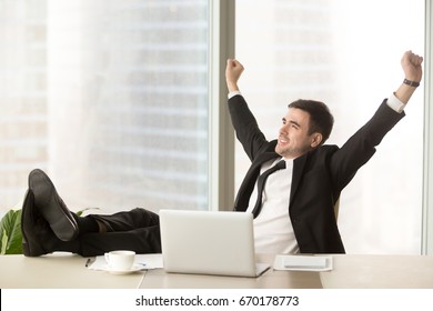 Satisfied businessman happy to finish work with laptop at office, raises hands and puts feet up on table, relaxing after hard working day in expectation of weekend leave, relaxed workday, no stress 