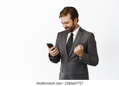 Satisfied businessman looking pleased at mobile phone, rejoicing and saying yes, triumphing, checking stock market data, white background