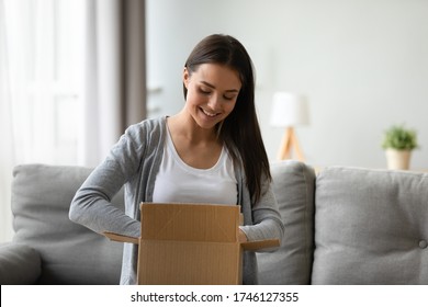 Satisfied client young beautiful woman seated on couch in living room holding cardboard on lap feels pleased of delivery service unpacking box smiling enjoy moment of unbox purchased goods in web shop