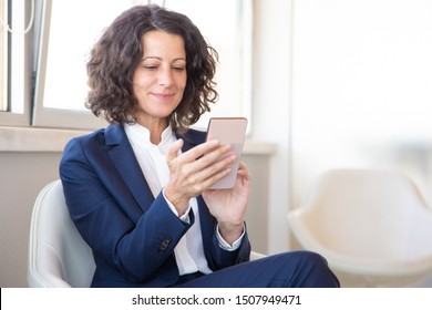 Satisfied Customer Using Online Mobile App. Business Woman Sitting In Armchair, Using Mobile Phone, Looking At Screen And Smiling. Digital Technology Concept