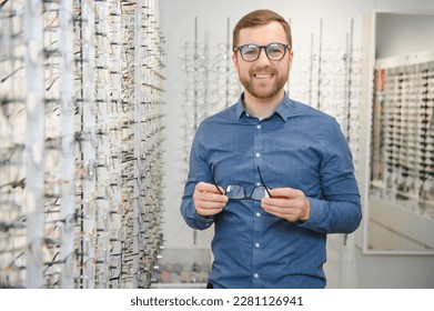 Satisfied Customer. View of happy young male client wearing new glasses, standing near rack and showcase with eyewear. Smiling man trying on spectacles.