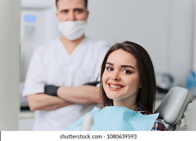 Satisfied dentist patient showing her perfect smile after treatment in a clinic box with dentist in the background