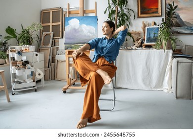 Satisfied dreaming artist woman resting on chair in art studio, smiling looks aside, pondering creative ideas for new pictures. Contented relaxed young female painter take break from painting process. - Powered by Shutterstock