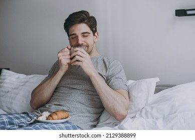 Satisfied Excited Young Man 20s In Pajamas Grey T-shirt Lying Sit In Bed Hold Cup Drink Coffee Eat Croissant Look Aside Resting Relax At Home Indoors Bedroom Good Mood Night Morning Bedtime Concept.