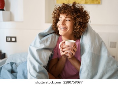 Satisfied female with cup of hot drink looking away while resting on bed in morning at home