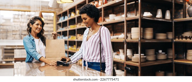 Satisfied female customer making a contactless NFC payment using her smartphone. Cheerful young woman smiling happily while shopping from a local female-owned ceramic store. - Powered by Shutterstock