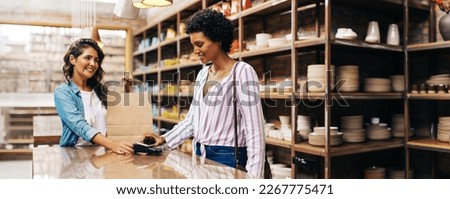 Satisfied female customer making a contactless NFC payment using her smartphone. Cheerful young woman smiling happily while shopping from a local female-owned ceramic store.