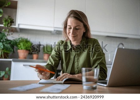 Satisfied female engaged financial receipts at home writing down expenses in diary notebook, paying utilities using laptop. Pleased woman planning family budget happy to find chance to save money.