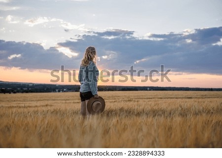 Satisfied female farmer is looking at barley field in cultivated land. Woman with straw hat standing in agricultural field during sunset