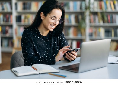 Satisfied female freelancer sits at the table in the office, using smartphone. Attractive woman browsing internet, taking a break from work, chatting with friends or family, smiling