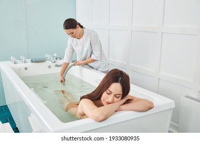 Satisfied Female Relaxing With Head On A Bathtub Rim With Spa Worker Taking Care Of Her Legs With Water Massage