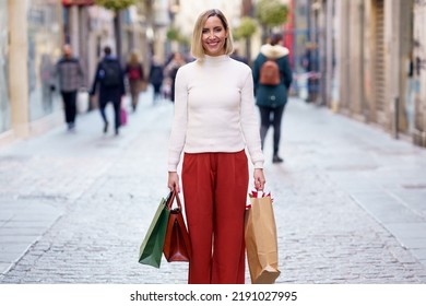 Satisfied female shopper in white turtleneck and pants standing with shopping packages and handbag on paved street and smiling at camera - Shutterstock ID 2191027995