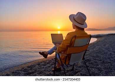 Satisfied free millennial freelancer woman using computer and sitting on beach by calm sea at sunset. Enjoyment of dream office remote work concept - Shutterstock ID 2147204403