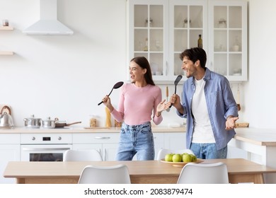 Satisfied Funny Attractive Millennial European Couple Have Fun, Singing At Imaginary Microphone, Preparing Food In Kitchen Interior. Joy And Cooking Together, Enjoy Weekend And Free Time, Copy Space