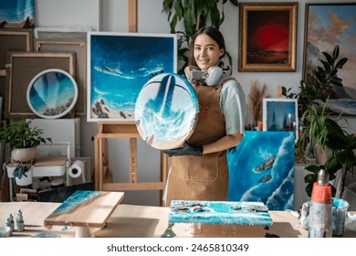 Satisfied girl artist holds finished epoxy resin painting, look to camera show off work in small business for sale. Sea picture of liquid malleable materials on canvas. Painter woman in art studio. - Powered by Shutterstock