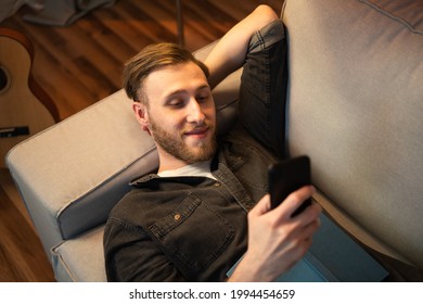 Satisfied Guy Is Lying On A Bright Couch Wearing A Black Shirt. One Hand Rests Under His Head Resting On The Headrest, The Other Holds His Phone And Texts His Friends, Arranges A Party