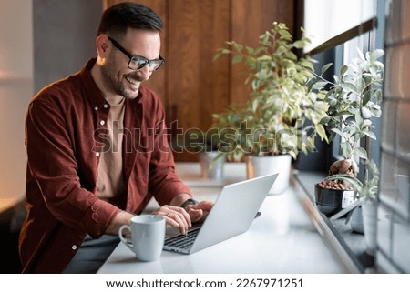 Satisfied handsome millennial man typing on laptop computer while sitting at table near window with potted plants, communicating online writing emails distantly working or studying on computer at home