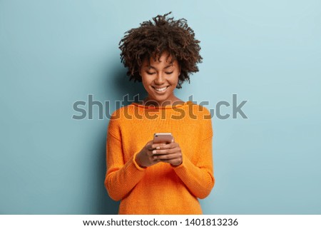 Satisfied hipster girl with Afro haircut, types text message on cell phone, enjoys online communication, types feedback, wears orange jumper, isolated on blue studio wall. Technology concept