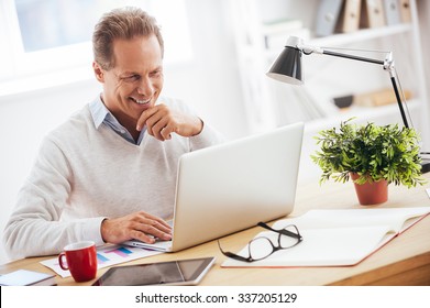 Satisfied With His Work. Cheerful Mature Man Working On Laptop And Smiling While Sitting At His Working Place