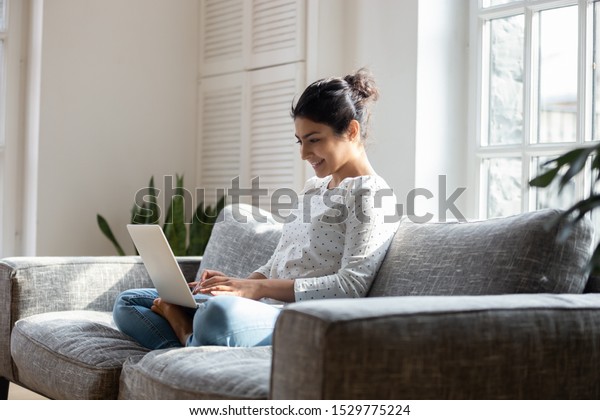 Satisfied Indian woman relaxing on comfortable\
couch, using laptop at home, happy girl chatting with friends in\
social network, spending lazy weekend, watching movie, shopping\
online, writing email