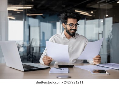 Satisfied Indian young man businessman working with documents, sitting in the office at the table and looking through the papers. - Powered by Shutterstock